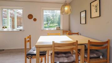 Un espace repas comprenant une table et des chaises en bois, un luminaire suspendu et deux fenêtres donnant sur un paysage verdoyant.