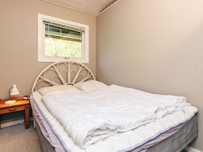 Una cama bien hecha con cabecera, una mesita de noche con lámpara, y una ventana sobre la cama.