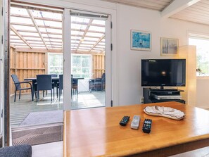 Une pièce avec une table en bois, un téléviseur et trois télécommandes. Une porte-fenêtre coulissante donne sur un espace extérieur aménagé avec une table et des chaises.
