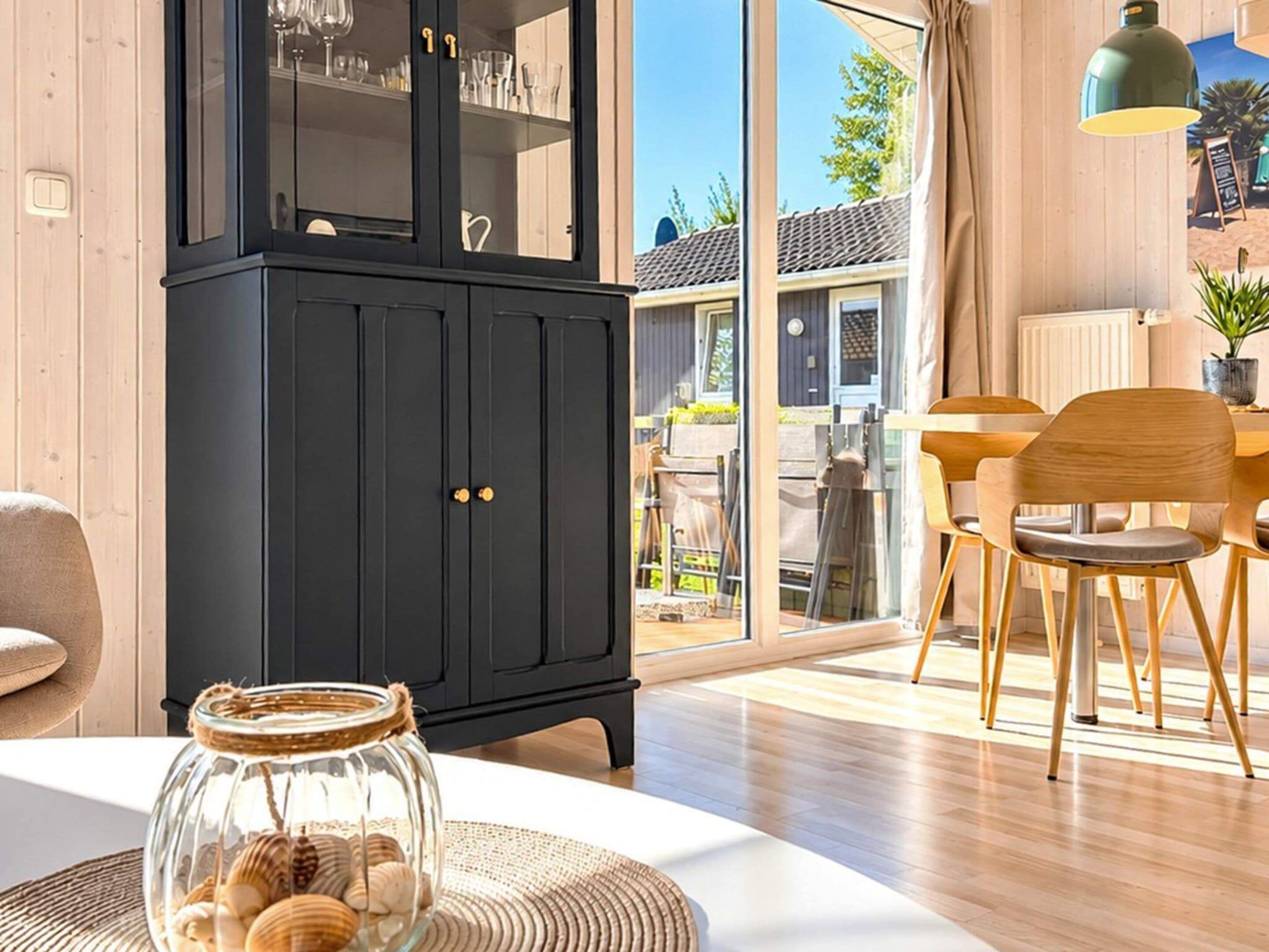 Een modern interieur met een zwarte kast, een eethoek met houten stoelen en een glazen pot met schelpen op een tafel.