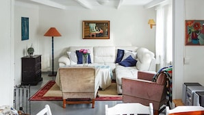 Un salon avec un canapé blanc, un petit fauteuil, une table basse et une table à manger avec des chaises. Il y a un tableau au mur, une lampe et un meuble sur lequel est posée une plante.
