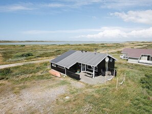 A single-story cabin with a porch, a small deck, and a satellite dish, surrounded by a grassy landscape and a body of water. - 6 Person Holiday Home in Lemvig (Lemvig)