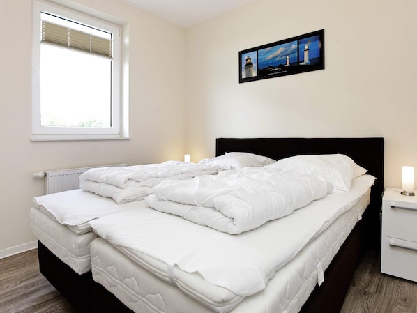 A double bed with white bedding, a nightstand with a lamp, a window with blinds, and a framed picture of lighthouses on the wall. - Holiday Home in Grossenbrode (Grossenbrode)