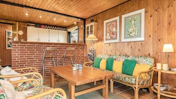 Una habitación acogedora con techo de madera, pared de ladrillo, una mesa de madera, muebles de mimbre y un sofá con cojines de colores.