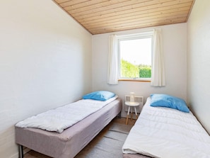 Un petit logement composé d’une seule pièce, avec deux lits simples, une fenêtre avec des rideaux, une petite table et un radiateur.