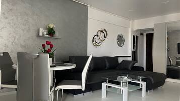 Ein modernes Wohnzimmer mit einem grauen Sofa, einem Glastisch und einem Essbereich mit weißem Tisch und Stühlen.