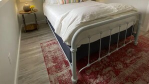A bedroom with a white metal bed frame, a white bedspread, and a striped pillow. There are two bedside tables with lamps, a red and white patterned rug, and a window on the left.