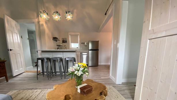 A dining area with a wooden table and chairs, a vase with flowers, and a kitchen in the background.