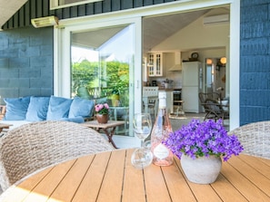 An outdoor seating area with wicker furniture, a table with a vase of purple flowers, a bottle of Sandara sparkling wine, and a glass.