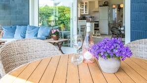 Ein Außenbereich mit Korbmöbeln, einem Tisch mit einer Vase voller lila Blumen, einer Flasche Sekt und einem Glas.