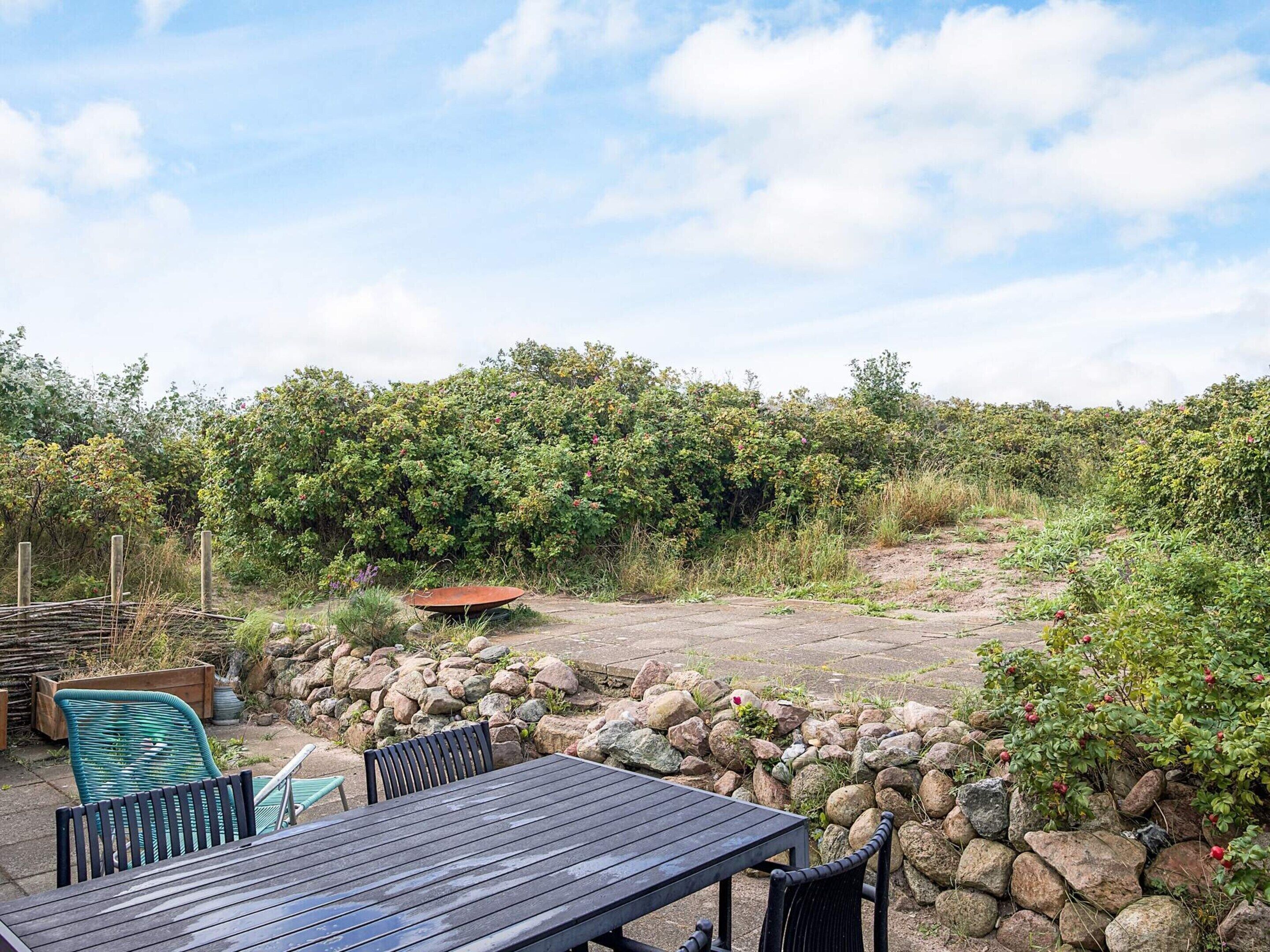 Un espace extérieur aménagé en terrasse, avec une table et des chaises en bois, entouré d’un mur de pierre et d’une végétation luxuriante.