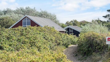 A coastal building with a red exterior and a metal roof, surrounded by dense greenery and a sandy path leading to it.