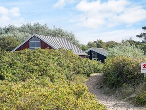 A coastal building with a red exterior and a metal roof, surrounded by dense greenery and a sandy path leading to it. - 6 Person Holiday Home in Store Fuglede (Store Fuglede)
