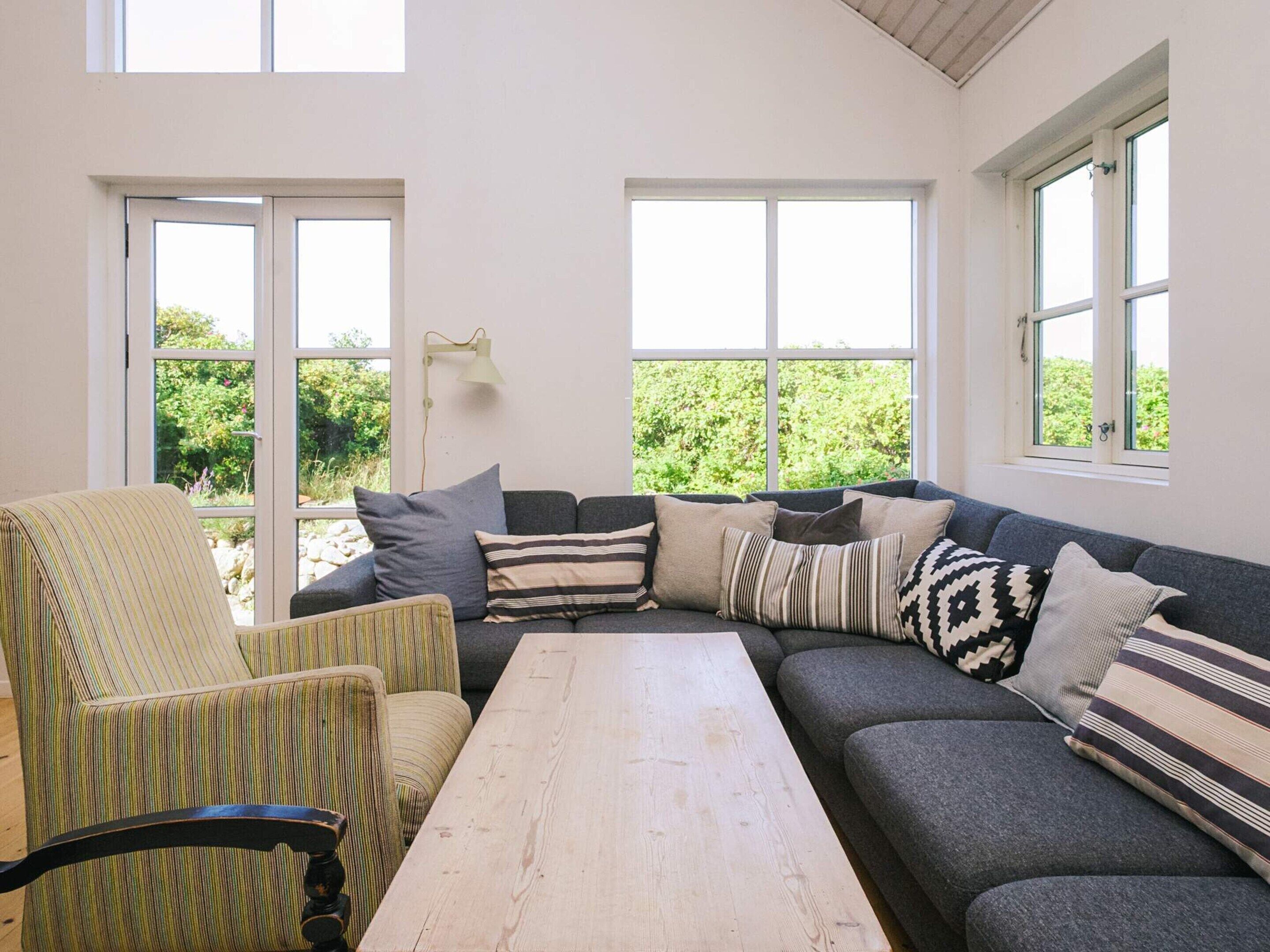 A cozy living room with a sofa, a chair, and a table. There are large windows allowing natural light and views of greenery outside.