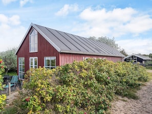 A red barn-style building with a sloped roof, surrounded by dense greenery and a sandy path. - 6 Person Holiday Home in Store Fuglede (Store Fuglede)