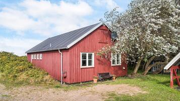 A red wooden house with a sloped roof, a bench, and a tree with white blossoms.