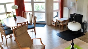 A room with a dining table, chairs, a sofa, a small table with a bottle of wine, and a hanging light fixture. - Holiday Home in Otterndorf (Otterndorf)