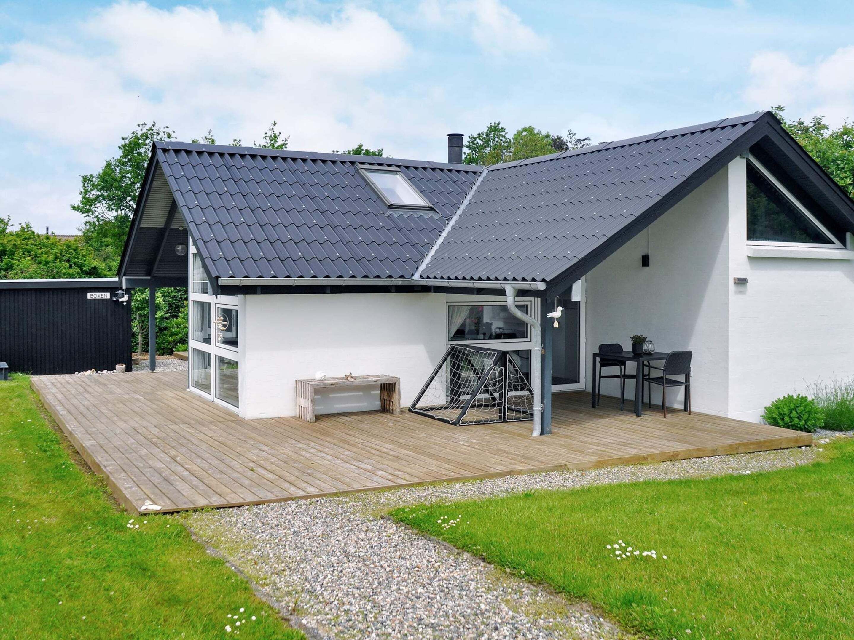 A modern house with a sloped roof, a wooden deck, a small outdoor seating area, and a large window.