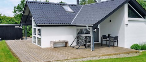 A modern house with a sloped roof, a wooden deck, a small outdoor seating area, and a large window.