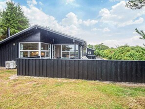 A black wooden cabin with large windows, a covered patio, and a visible air conditioning unit. - 5 Star Holiday Home in Henne (Henne)