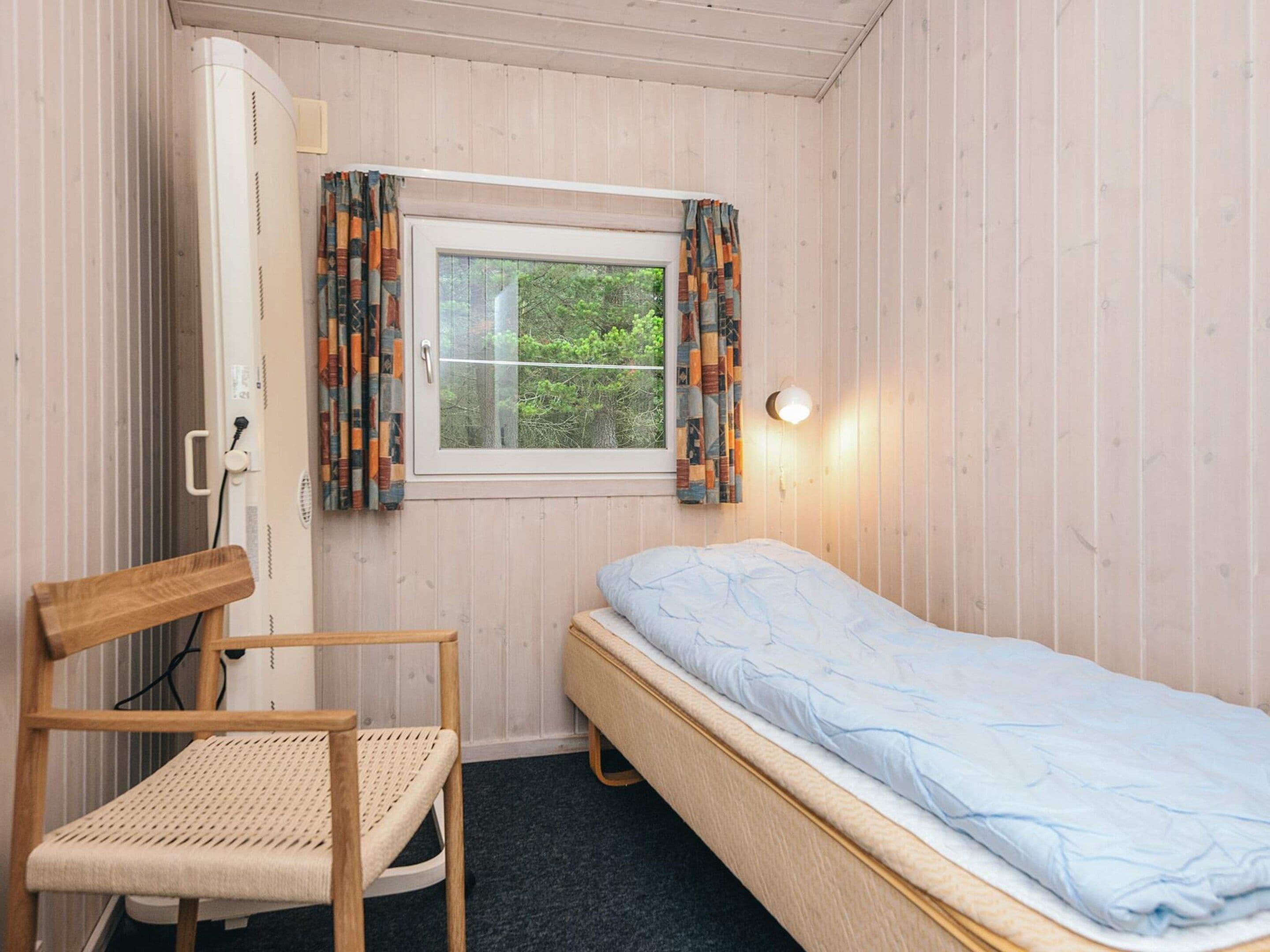 A small room with a single bed, a chair, a window with curtains, and a wall-mounted light.