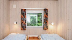 Ett rum med två enkelsängar, ett litet träbord, ett fönster med gardiner och två väggmonterade lampor.