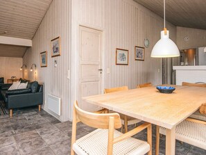 A cozy living room with a wooden dining table, chairs, and a sofa. There are framed pictures on the wall and a white door.