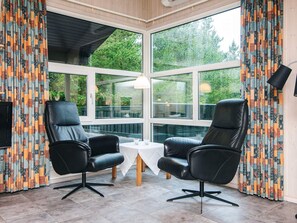 Quarto com duas poltronas de couro preto, uma mesa pequena branca e janelas amplas com cortinas estampadas.