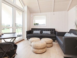 A modern living room with a dark leather sofa, round woven ottomans, and a large window providing a view of the outdoors. - 5 Star Holiday Home in Vaeggerlose (Vaeggerlose)