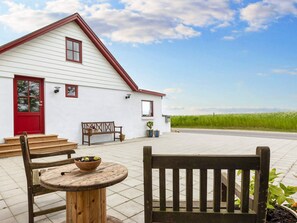 A white building with a red door and a red roof, a wooden bench, a small round table with a bowl, and potted plants.