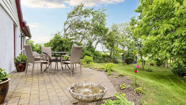 Une terrasse extérieure avec une table et des chaises, entourée de verdure et agrémentée d’un abreuvoir à oiseaux en pierre.