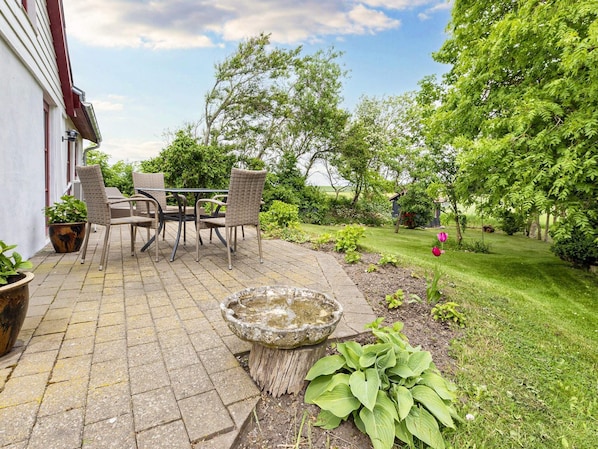 An outdoor patio with a table and chairs, surrounded by greenery and a stone birdbath.