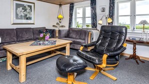 Un salon comprenant un canapé, une table basse en bois, un fauteuil en cuir noir et une vue sur l’extérieur grâce à de grandes fenêtres.