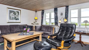 Uma sala de estar com sofá, mesa de centro de madeira, poltrona de couro preto e vista para o exterior através de grandes janelas.