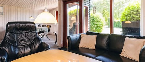 A modern interior with a wooden ceiling, a leather chair, a black sofa with cushions, a wooden table with two glasses, and a view through large glass doors.
