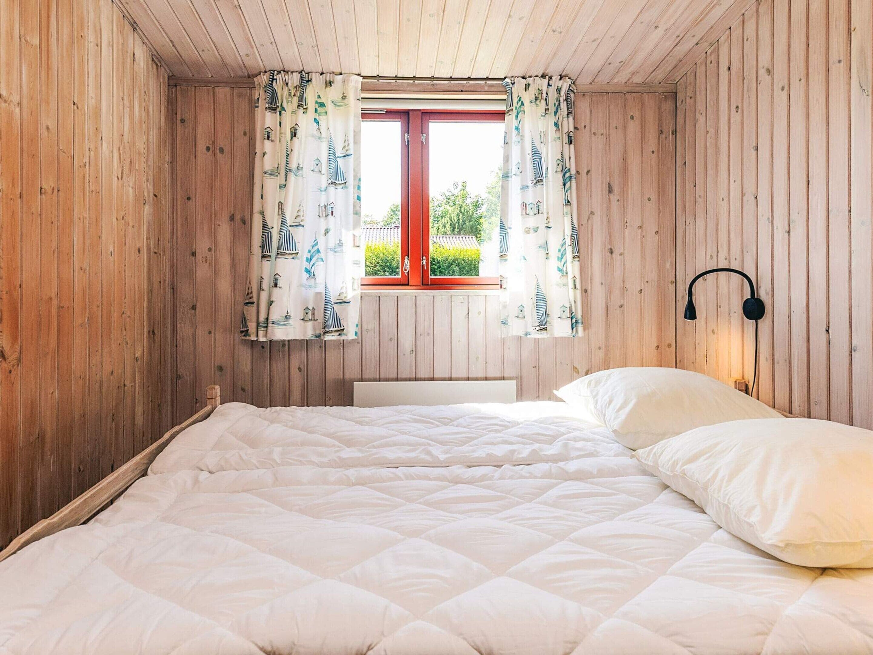 A bedroom with a bed, wooden walls, a window with curtains, and a wall-mounted lamp.
