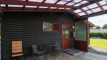 A covered porch with a wooden deck, a red door, and two chairs.