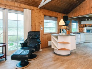 Un interior de cabaña acogedor con detalles de madera, un sillón de cuero negro, una pequeña cocina y un ventanal grande.