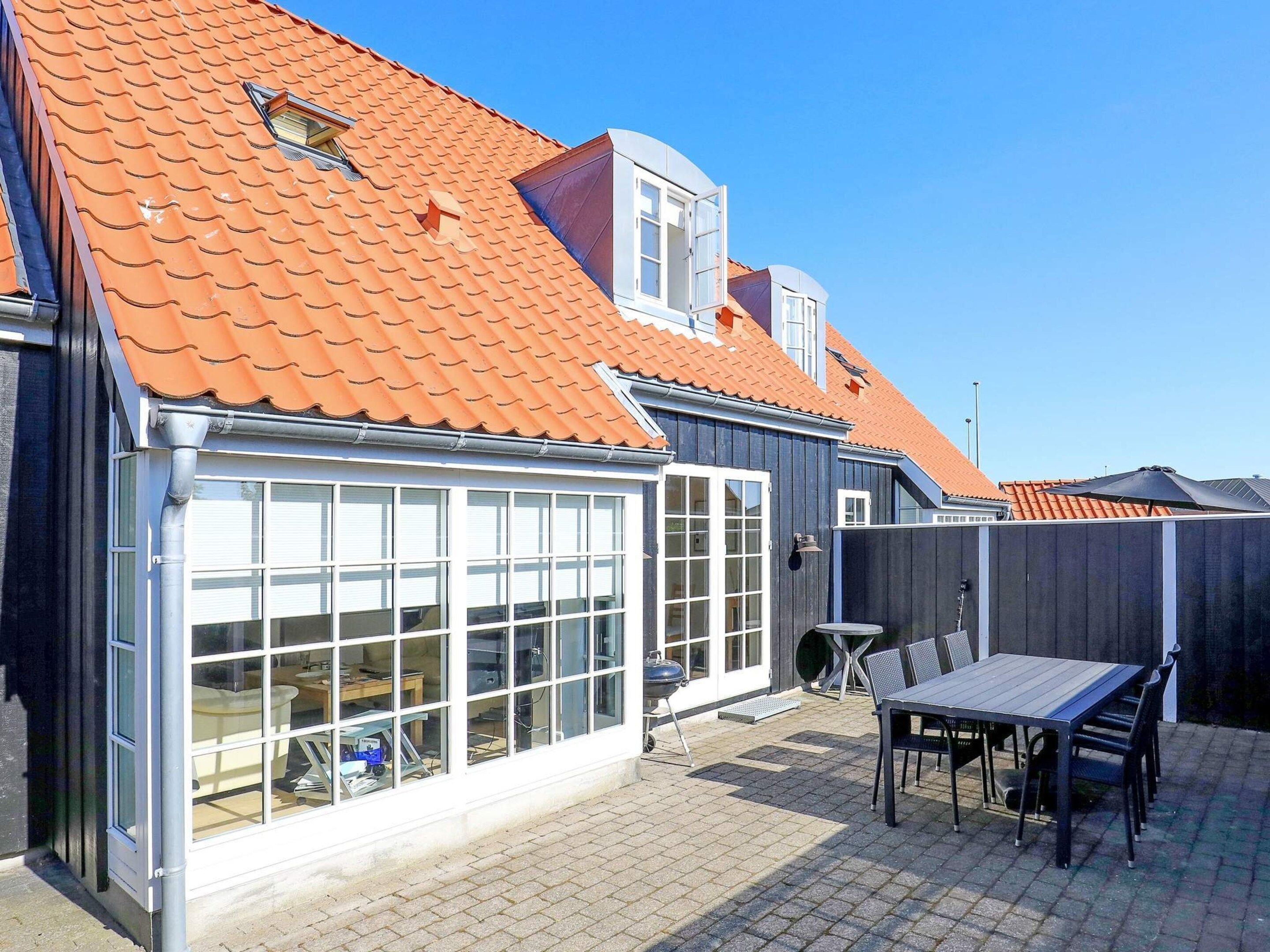 A residential building with a tiled roof, large windows, and a patio area with a table and chairs.