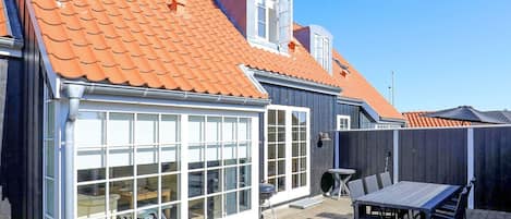 A residential building with a tiled roof, large windows, and a patio area with a table and chairs.
