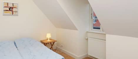 A bedroom with a sloped ceiling, a bed with a blue quilt, a bedside table with a lamp, and a window with a view of a red-roofed building.