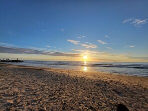 A beach with footprints leading to the water, a clear sky, and the sun setting on the horizon.