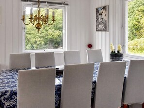 Une salle à manger agrémentée d’un lustre, de chaises blanches, d’une nappe à motifs et offrant une vue sur la verdure par la fenêtre.