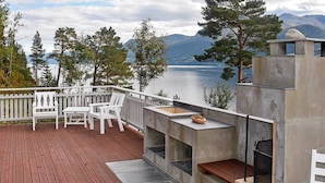 Eine Terrasse mit weißen Stühlen und einem Steinkamin mit Blick auf einen See, im Hintergrund Berge.