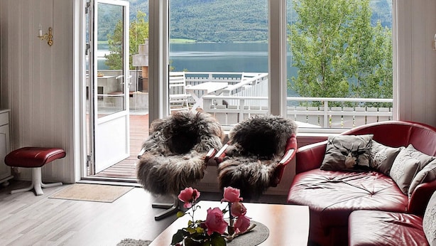 Ein Wohnzimmer mit Blick auf einen See und Berge, ein rotes Ledersofa, ein Couchtisch aus Holz mit einem Blumenstrauß und ein flauschiger Sessel.