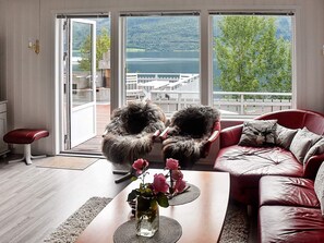 Un salon avec vue sur un lac et des montagnes, un canapé en cuir rouge, une table basse en bois ornée d’un vase de fleurs et un fauteuil moelleux.