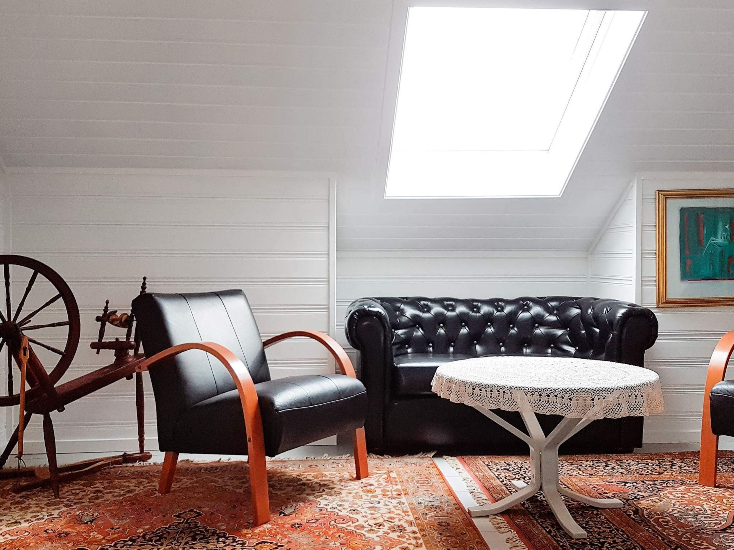 A loft-style room with a black leather sofa, a round table with a lace tablecloth, a vintage chair, and a large window.