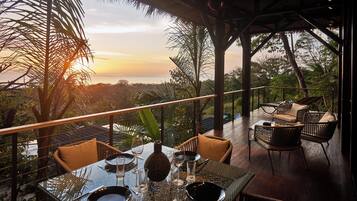 Un comedor con una mesa lista para dos, con vistas panorámicas del océano y palmeras al atardecer.