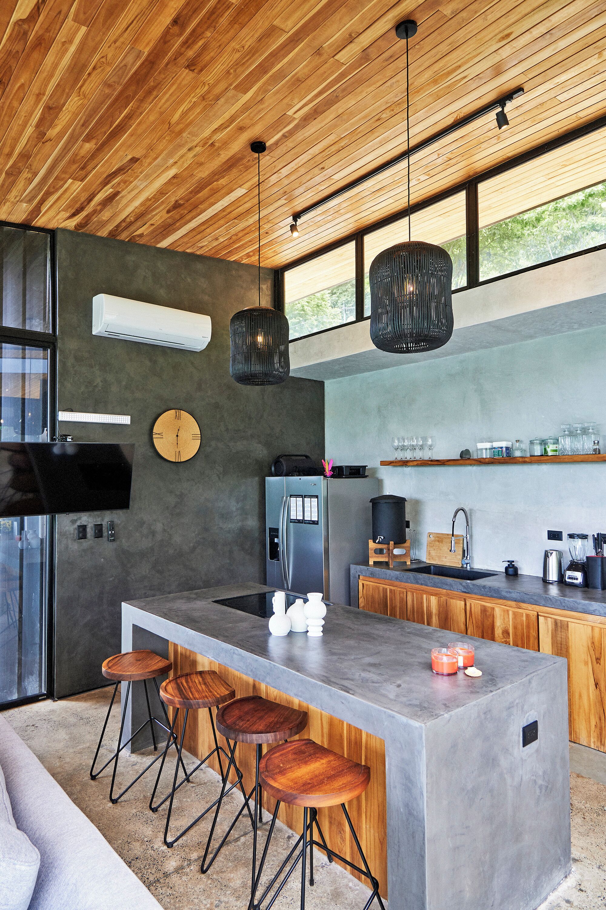 A modern kitchen with a large island, bar stools, wooden ceiling, and a large window.