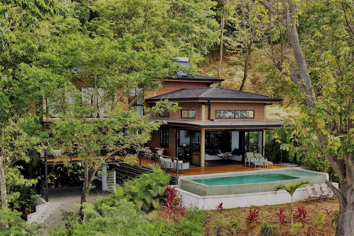Uma casa moderna com um deque espaçoso e uma piscina, cercada por vegetação exuberante.